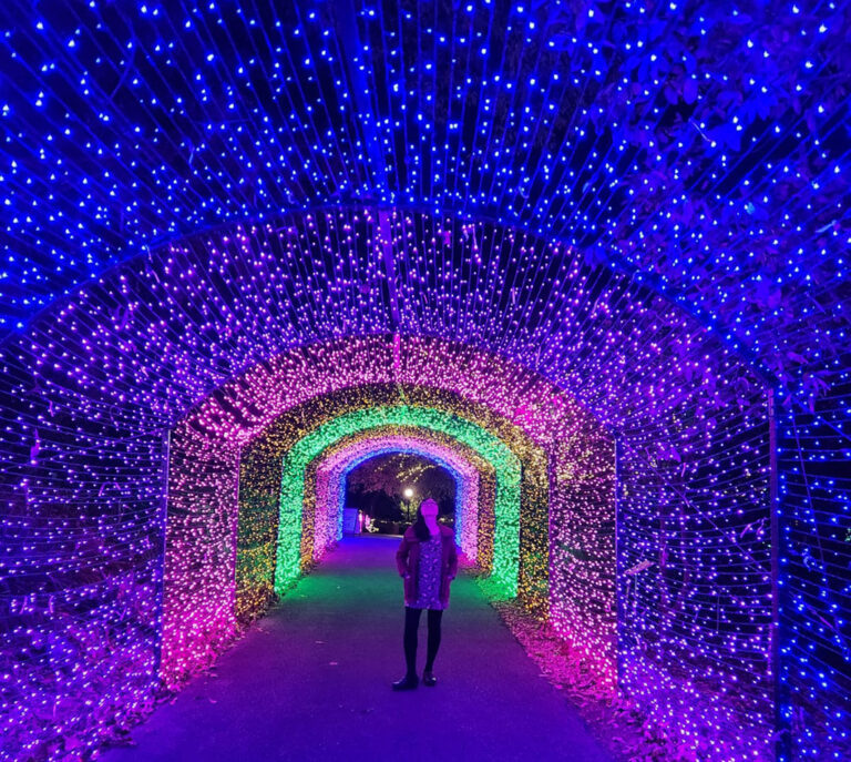 woman standing in a tunnel of rainbow colored christmas lights