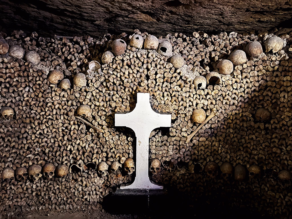 a cross gravestone in front of a wall of neatly stacked bones and arches of skulls