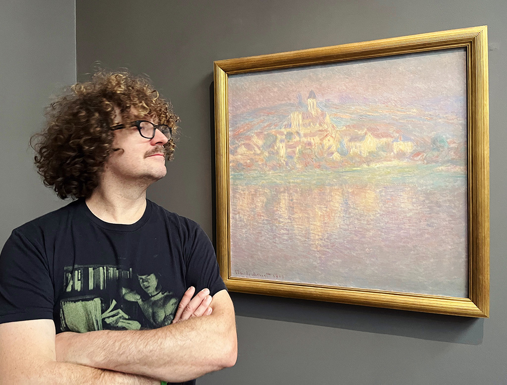 curly-haired man with cross-arms looking at painting of buildings reflected on water