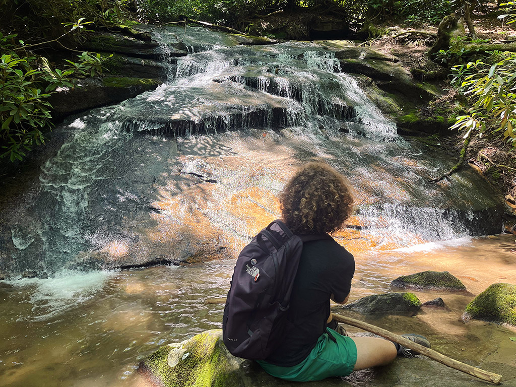 back of a man sitting on a rock looking out at a small waterfall