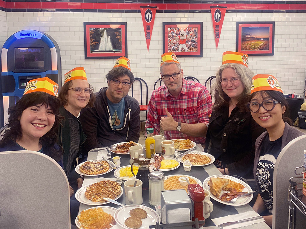 a group of friends gathered around a table full of breakfast food wearing Waffle House hats